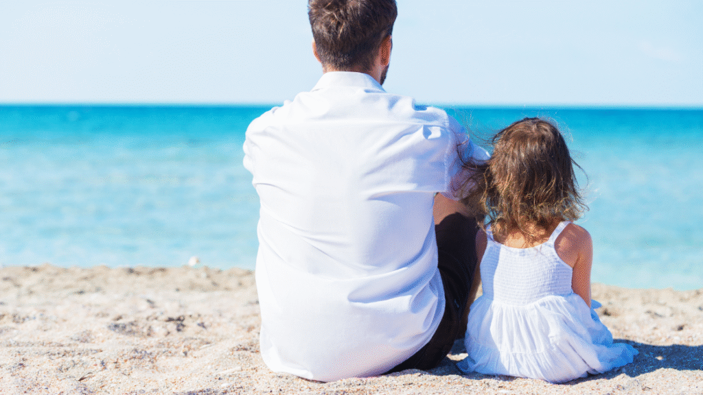 A man and a young girl sitting on the beach, looking out at the ocean.