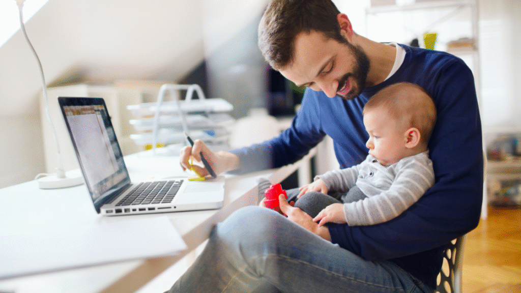 A man sitting at a desk with a laptop, holding a baby in his lap while writing notes.